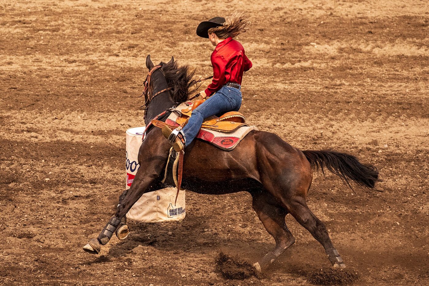 A cowgirl in a red shirt leaning into a barrel turn on a bay horse, hat brim hiding her face.
